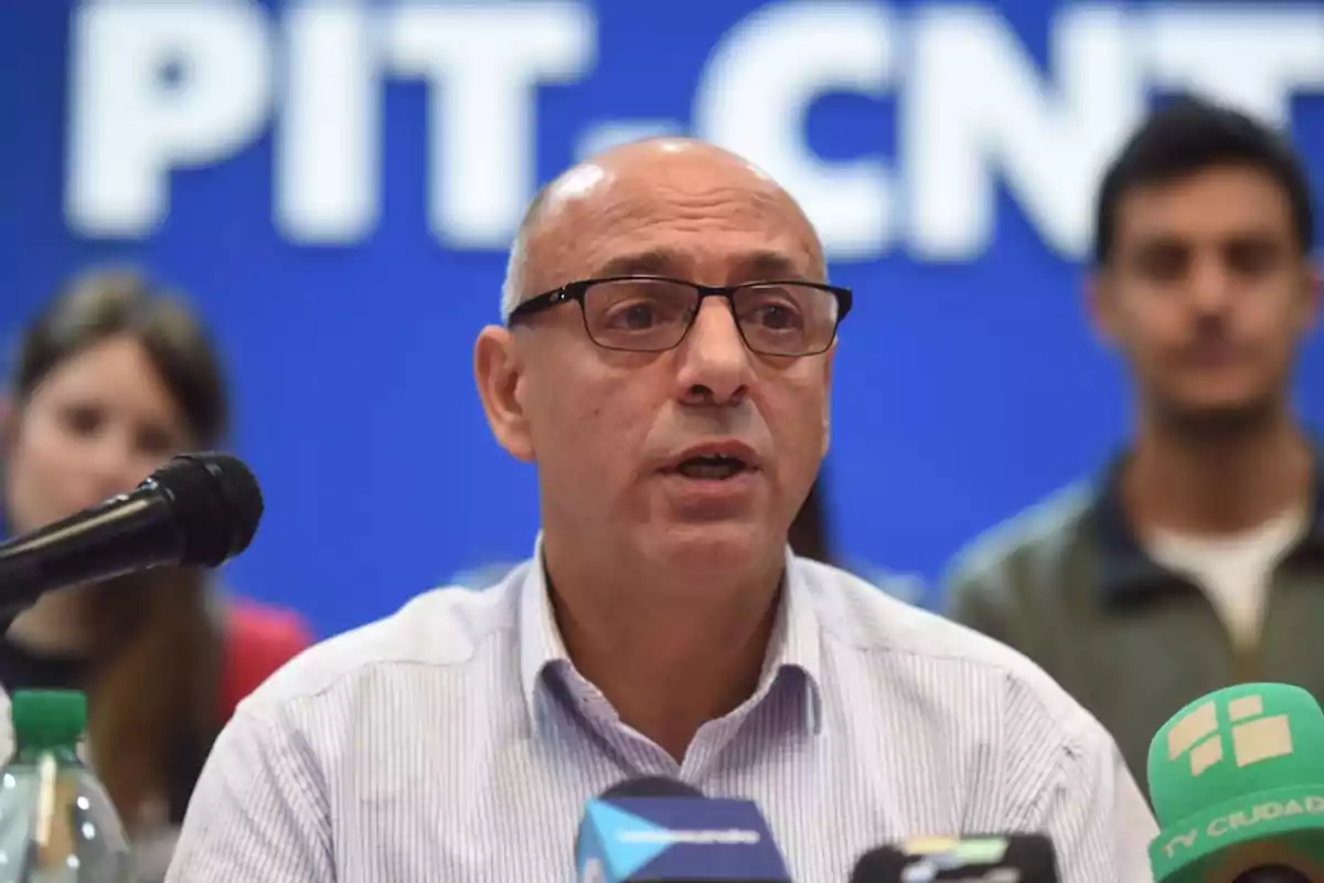 A man with glasses speaks in front of several microphones at a press conference with a blue background that has white letters.