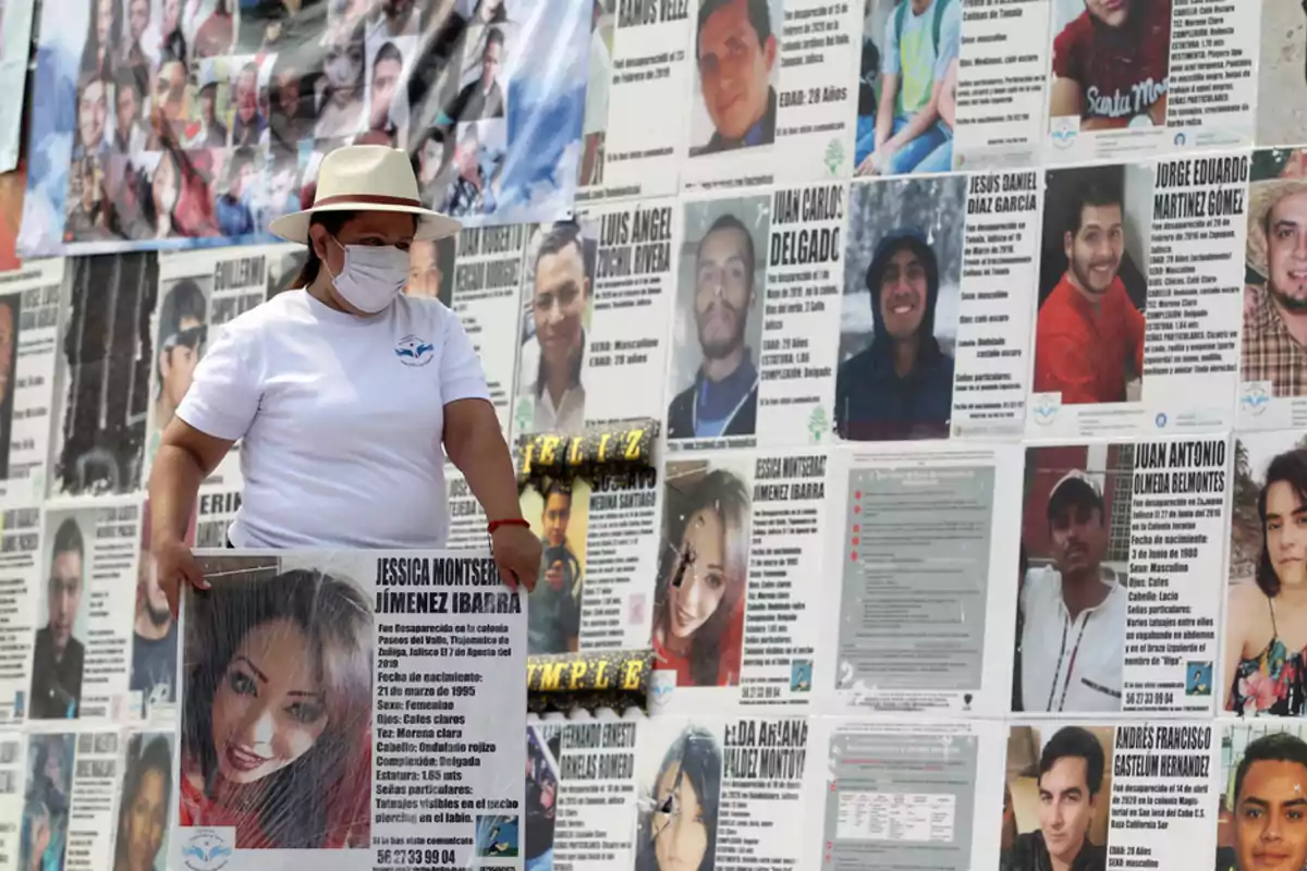 Una persona con sombrero y mascarilla sostiene un cartel con la foto de una mujer frente a un muro cubierto de carteles de personas desaparecidas.