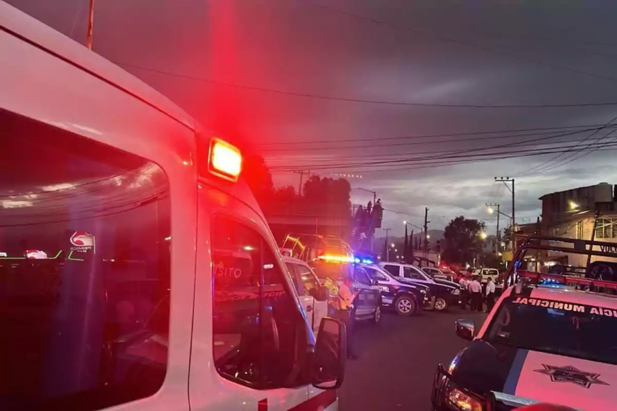 Una escena nocturna muestra vehículos de emergencia con luces encendidas en una calle, incluyendo una ambulancia y varios coches de policía.