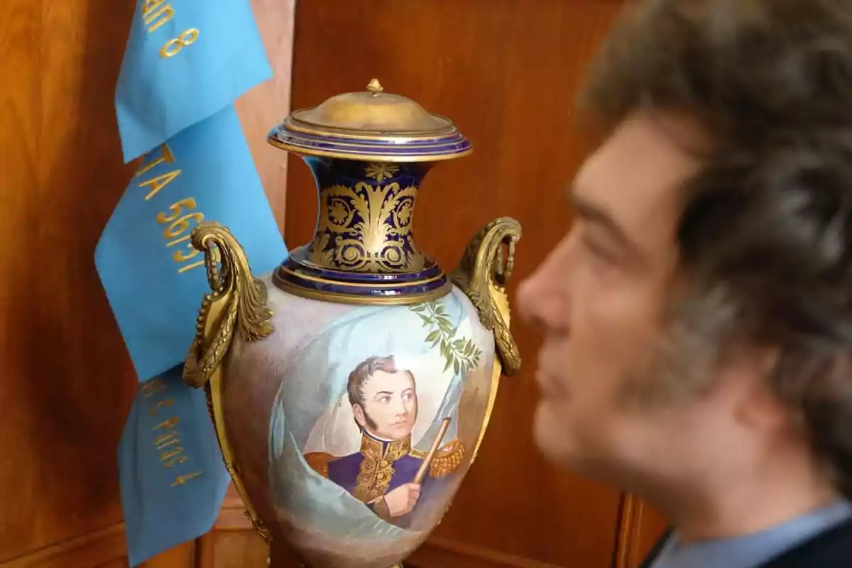 Un hombre de perfil observa un jarrón decorativo con un retrato de un militar, junto a una bandera azul con texto dorado. Un hombre de perfil observa un jarrón decorativo con un retrato de un militar, junto a una bandera azul con texto dorado.