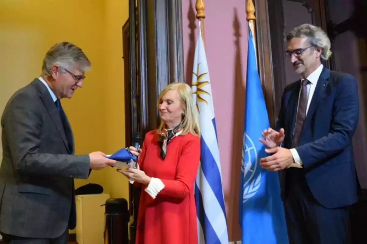 Three elegantly dressed people take part in a ceremony in front of the flags of Uruguay and the UN; a woman in the center holds a microphone and a blue object while the men beside her watch and applaud.
