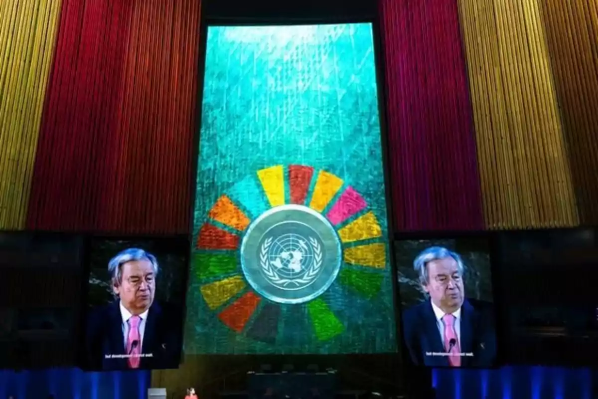 UN auditorium with a large screen displaying the Sustainable Development Goals logo and two side screens showing a gray-haired man in a pink tie speaking
