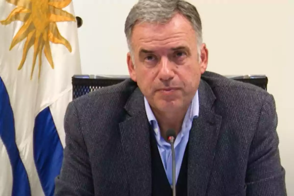 Gray-haired man in a gray jacket speaking in front of a microphone with a Uruguayan flag in the background