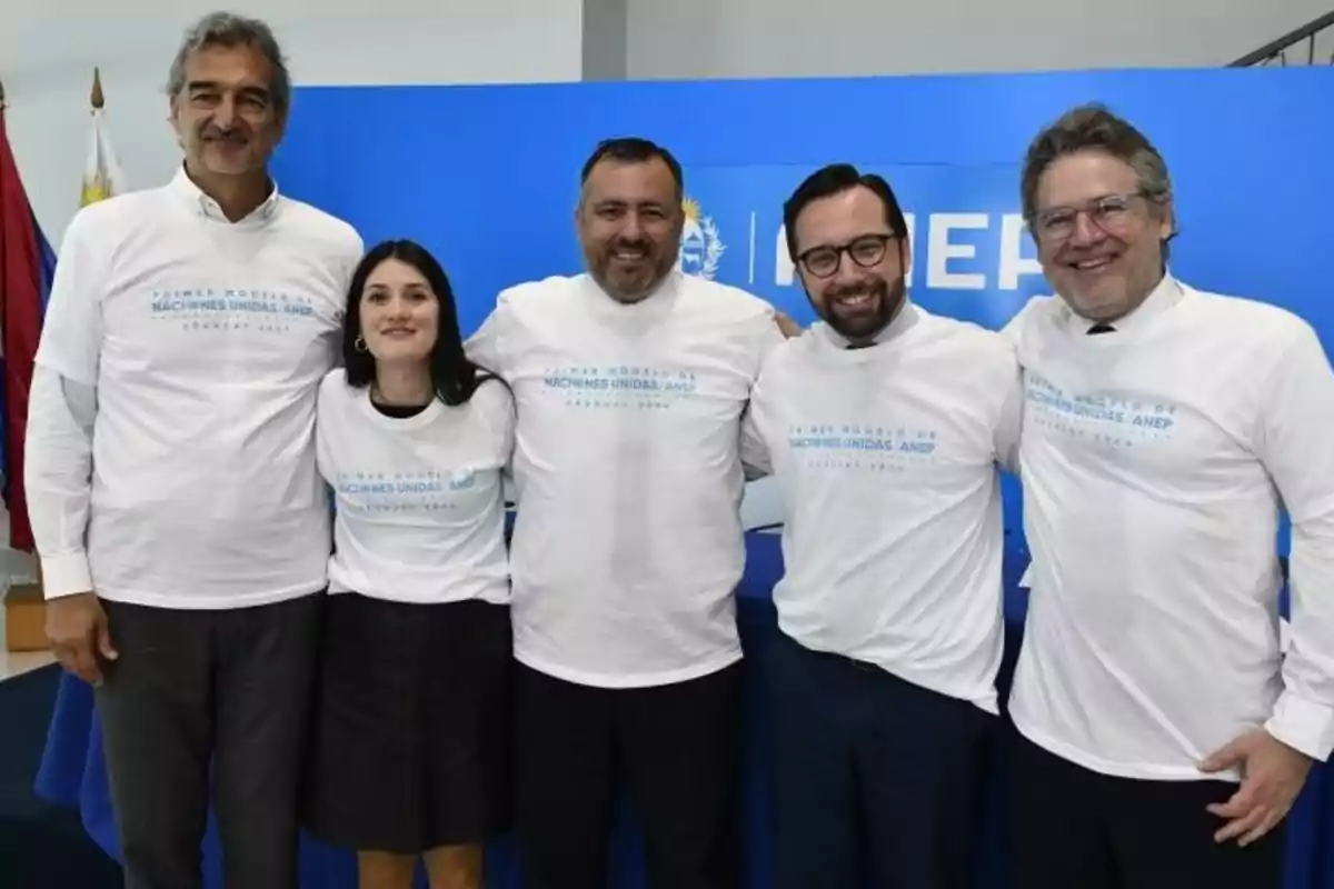Five people smile and pose together wearing white t-jerseys with blue text at an institutional event in front of a blue background.