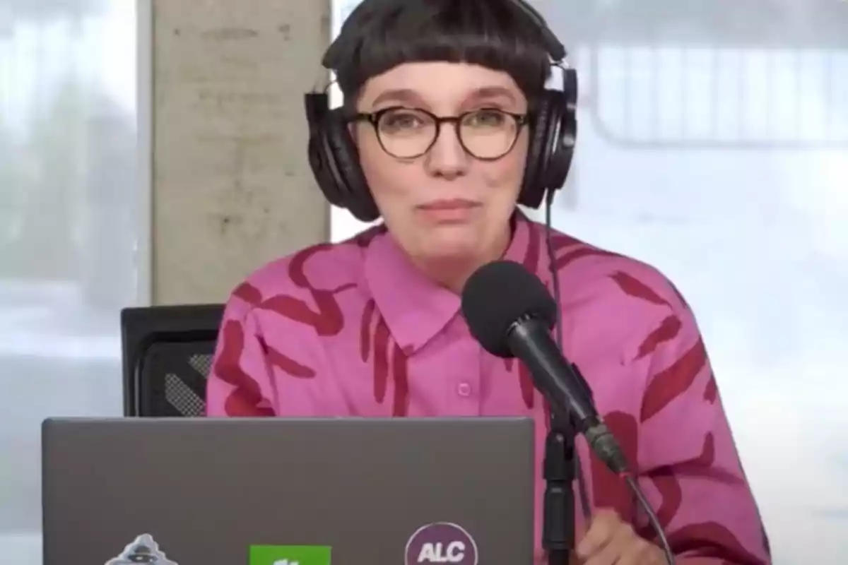 Person with glasses and large headphones in front of a microphone, wearing a pink jersey with a red print and a laptop with stickers