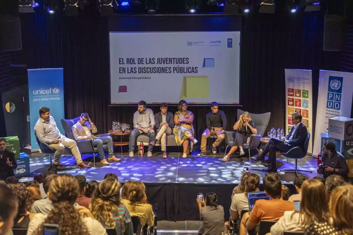 Un panel de jóvenes y adultos discute sobre el rol de las juventudes en las discusiones públicas en un auditorio con público y banners de UNICEF y la ONU al fondo