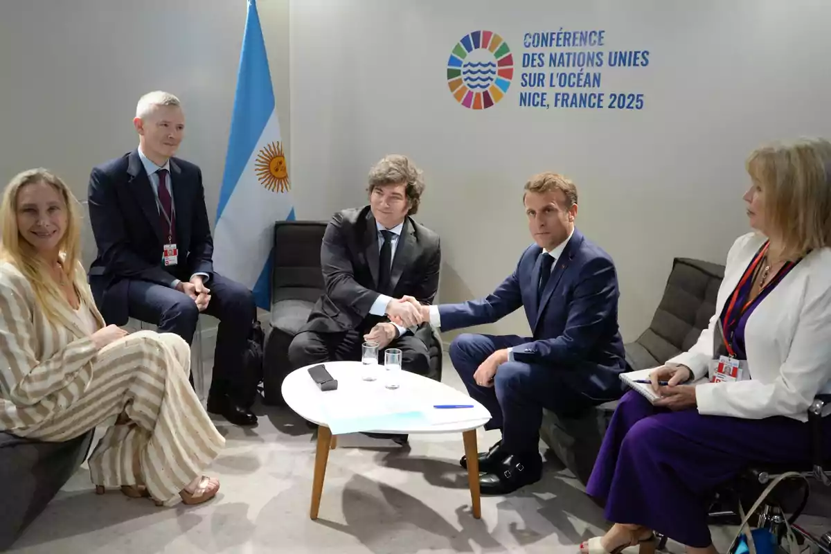 Cinco personas elegantemente vestidas se encuentran sentadas en una sala de reuniones, dos de ellas se dan la mano frente a una mesa pequeña, al fondo hay una bandera de Argentina y un cartel de la conferencia de la ONU sobre el océano en Niza, Francia 2025. Cinco personas elegantemente vestidas se encuentran sentadas en una sala de reuniones, dos de ellas se dan la mano frente a una mesa pequeña, al fondo hay una bandera de Argentina y un cartel de la conferencia de la ONU sobre el océano en Niza, Francia 2025.