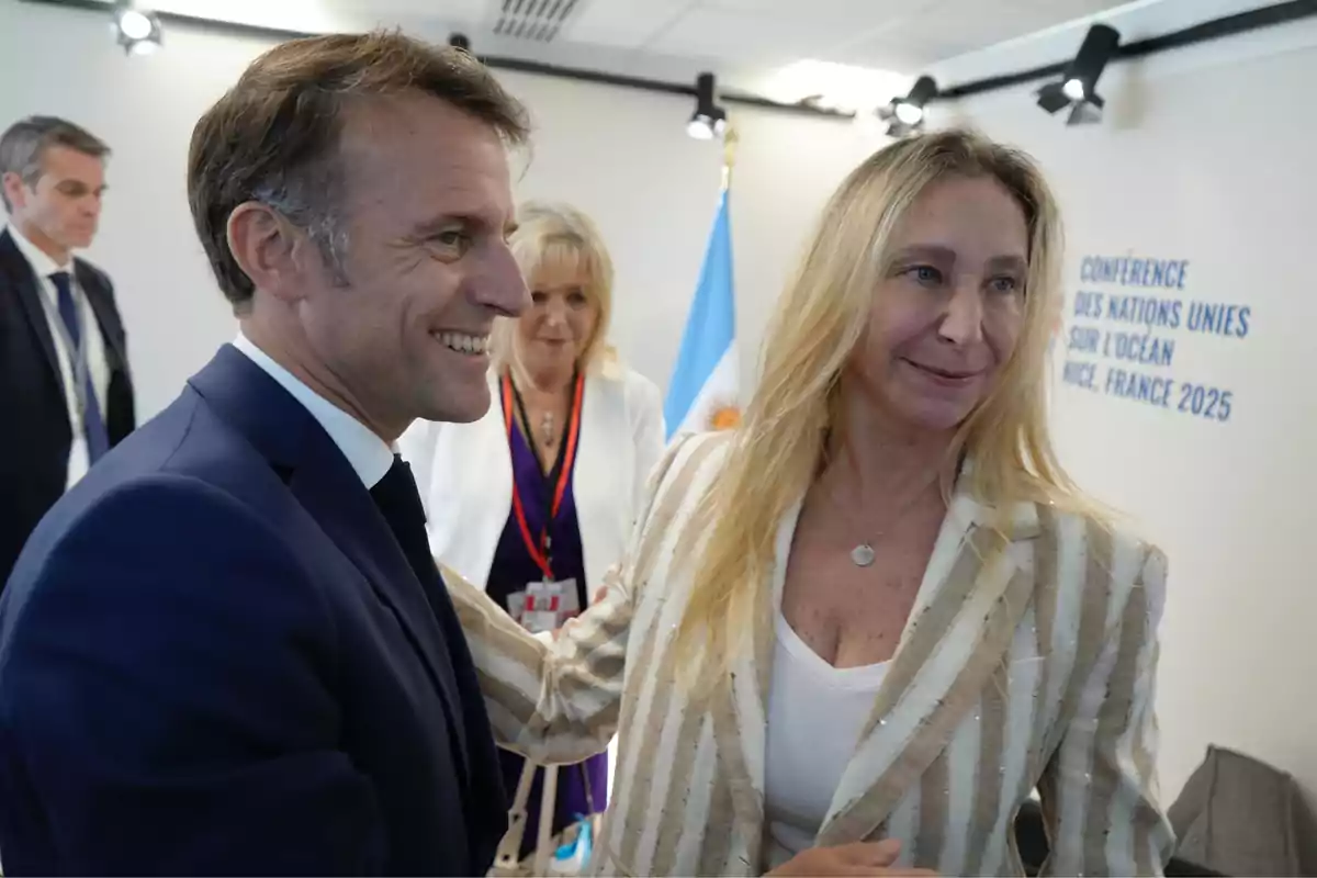 Un hombre con traje azul sonríe mientras saluda a una mujer rubia con saco a rayas en un evento internacional, al fondo se ve una bandera y personas conversando. Un hombre con traje azul sonríe mientras saluda a una mujer rubia con saco a rayas en un evento internacional, al fondo se ve una bandera y personas conversando.