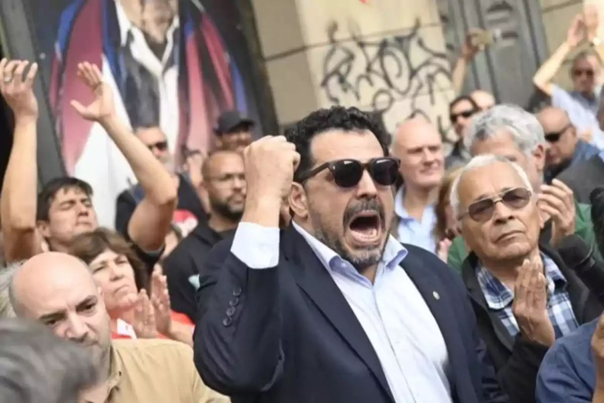 Man wearing sunglasses and a suit raising his fist and shouting at a protest, surrounded by people clapping and showing support Man wearing sunglasses and a suit raising his fist and shouting at a protest, surrounded by people clapping and showing support