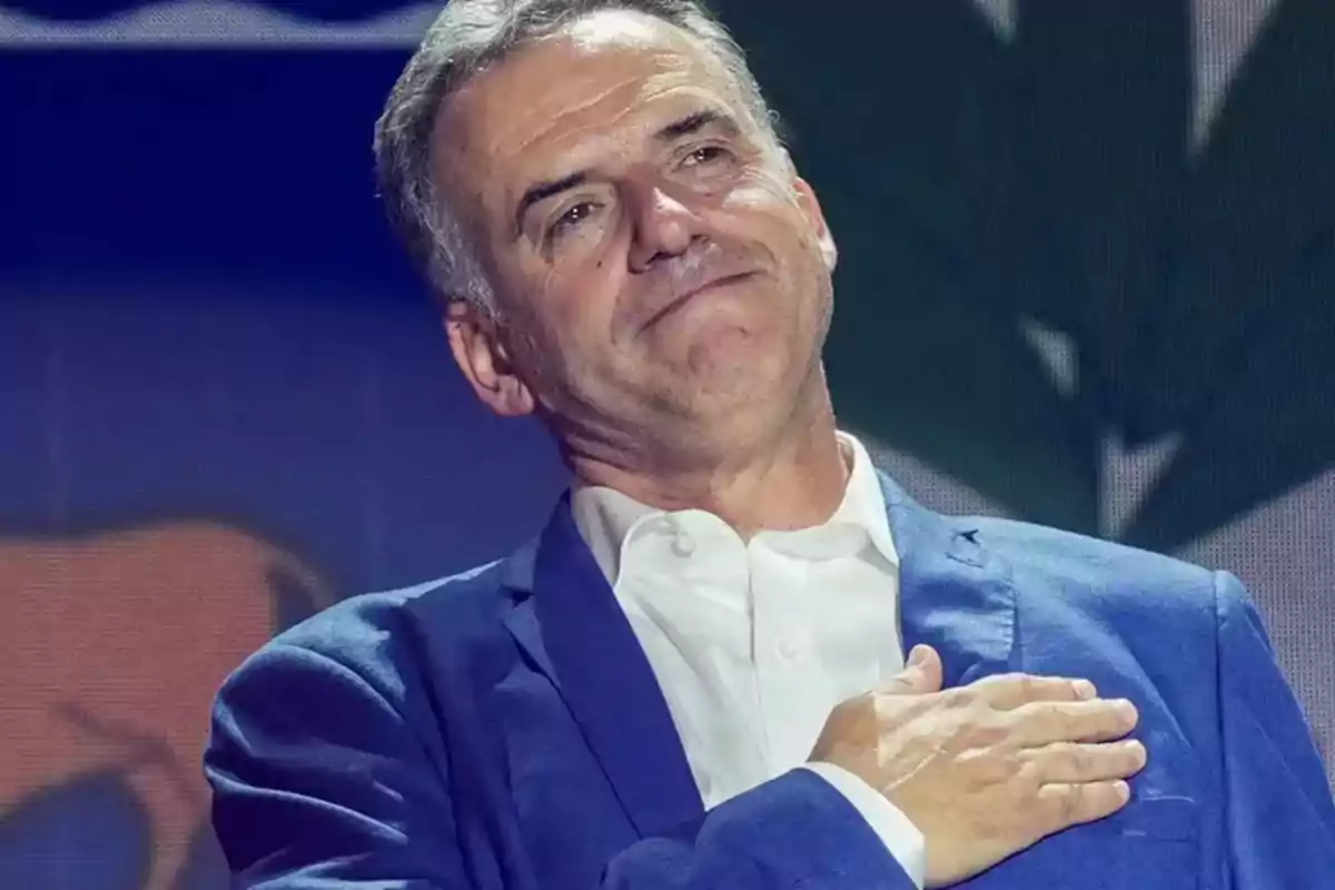 Gray-haired man with an emotional expression places his hand on his chest and wears a blue jacket and white jersey.