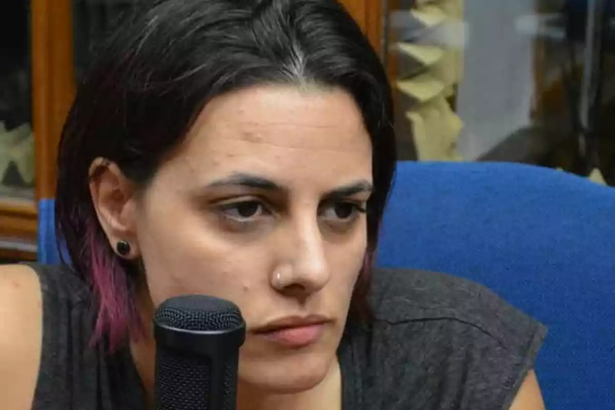 Dark-haired woman with short hair and purple highlights sitting in front of a microphone, with a serious expression Dark-haired woman with short hair and purple highlights sitting in front of a microphone, with a serious expression