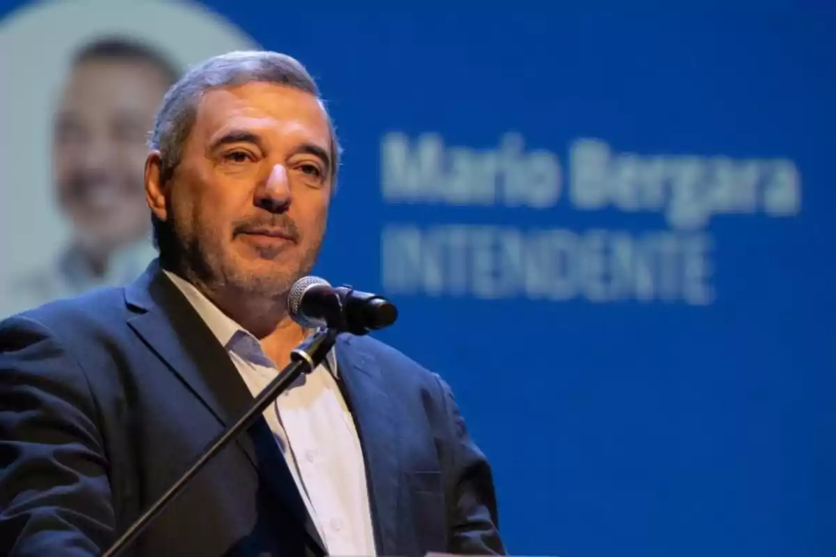 Hombre de cabello canoso y barba corta hablando frente a un micrófono con fondo azul donde se leen palabras desenfocadas Hombre de cabello canoso y barba corta hablando frente a un micrófono con fondo azul donde se leen palabras desenfocadas