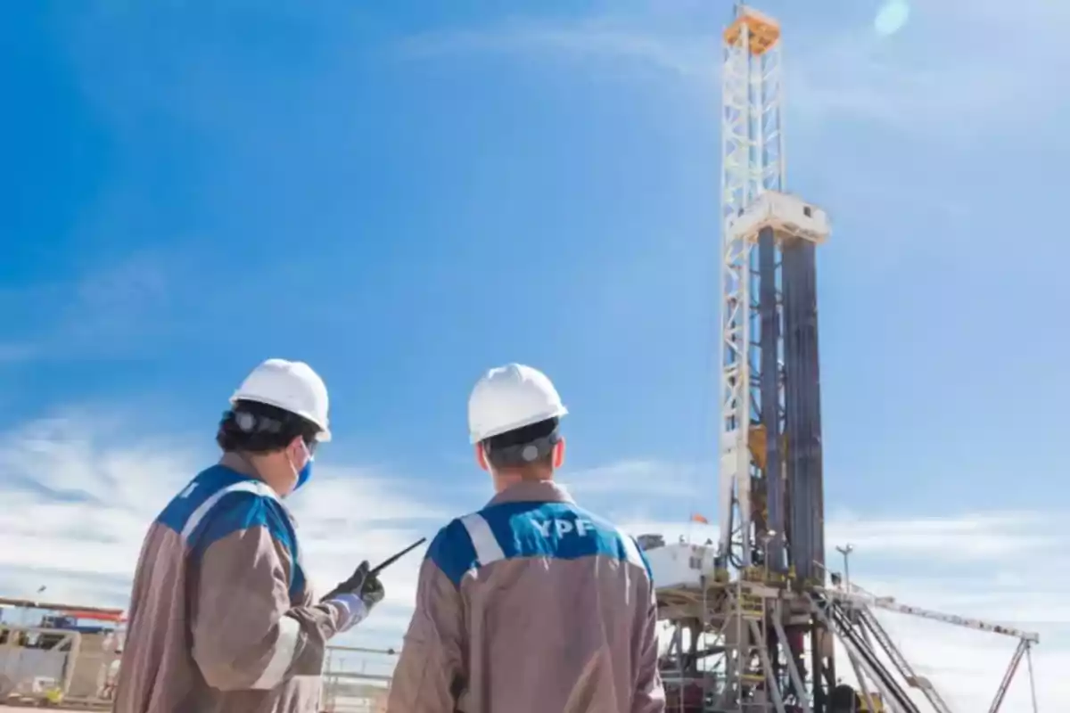 Two workers with white helmets observe a drilling tower at an industrial site under a clear sky.