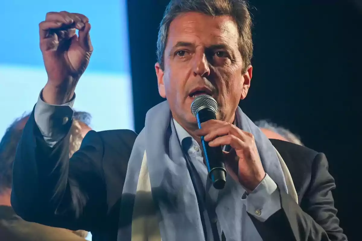 A man speaking into a microphone with an emphatic gesture and a light blue scarf over his shoulders. A man speaking into a microphone with an emphatic gesture and a light blue scarf over his shoulders.