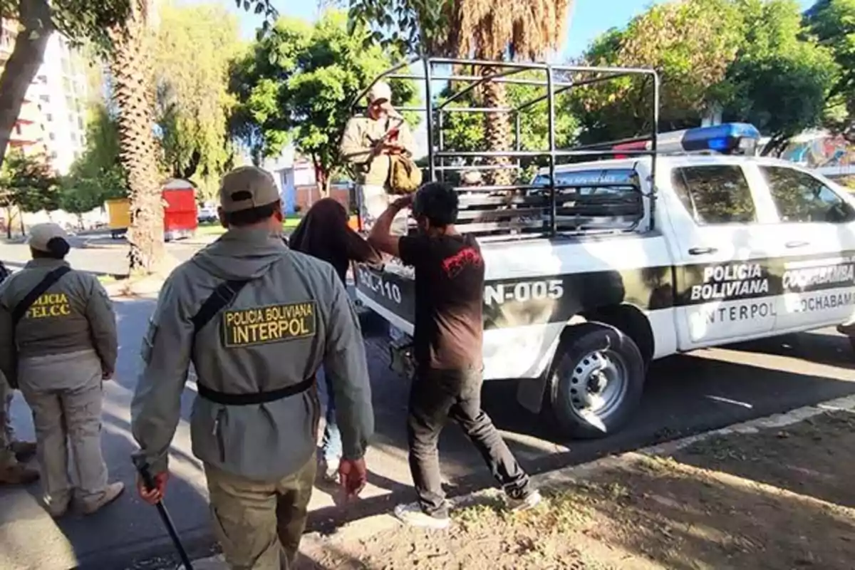 Un grupo de oficiales de la policía boliviana escolta a una persona hacia un vehículo policial en una calle arbolada.