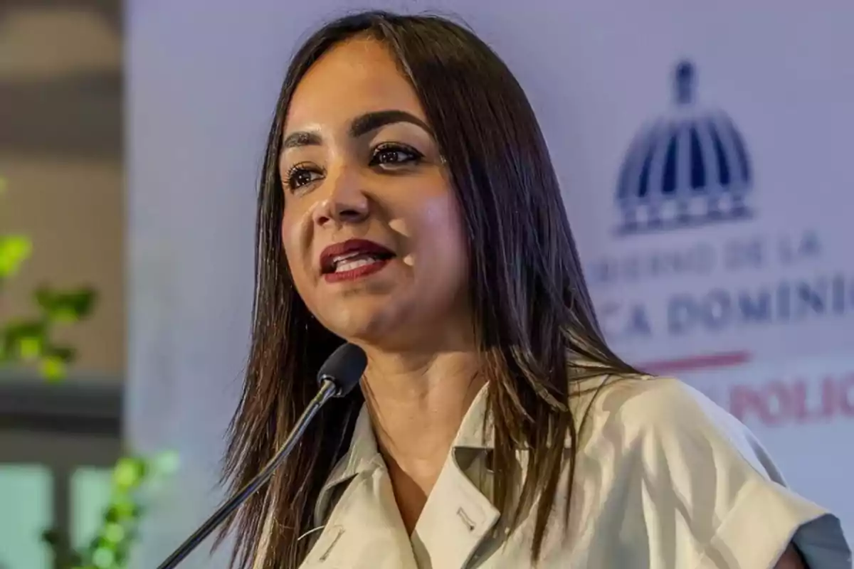 Una mujer hablando frente a un micrófono con un fondo que tiene un logotipo y texto borroso.