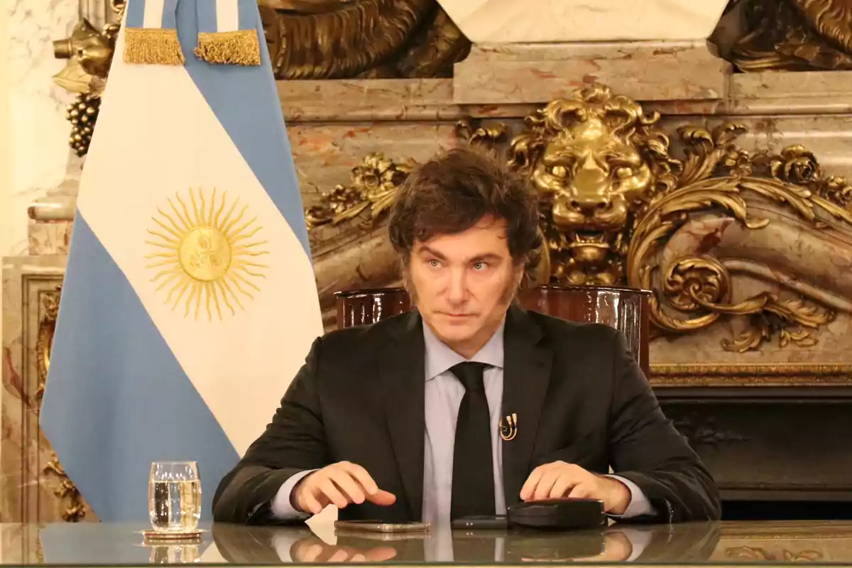 Un hombre con traje oscuro está sentado frente a una mesa con un vaso de agua y un micrófono, al fondo se ve una bandera de Argentina y una decoración dorada.