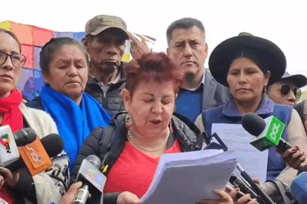 A group of people is gathered while a woman in the center reads a document in front of several press microphones.