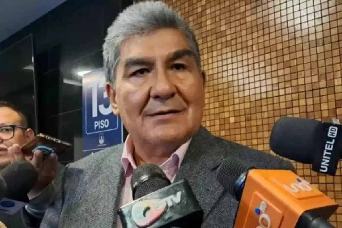 Un hombre de cabello canoso y traje gris está rodeado de micrófonos de diferentes medios de comunicación mientras parece estar dando una declaración en un lugar con un fondo de mosaico marrón y un cartel que indica el piso 13. Un hombre de cabello canoso y traje gris está rodeado de micrófonos de diferentes medios de comunicación mientras parece estar dando una declaración en un lugar con un fondo de mosaico marrón y un cartel que indica el piso 13.