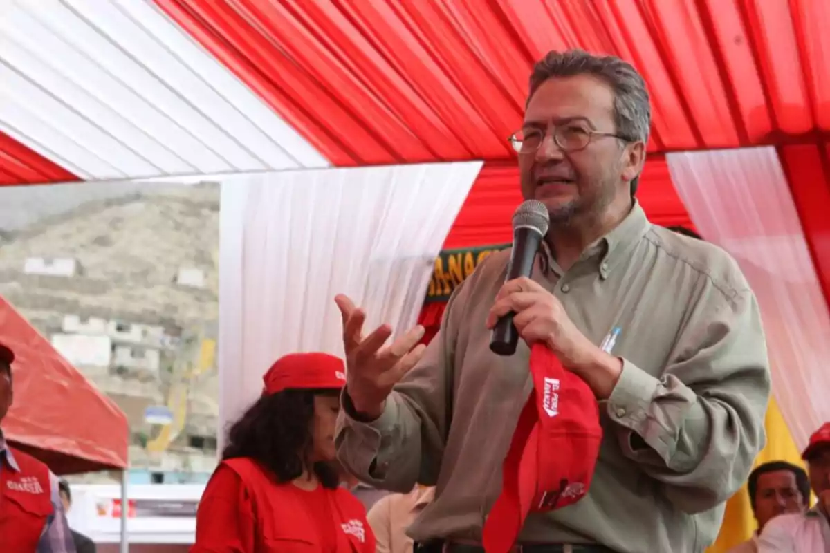 Un hombre con camisa verde sostiene un micrófono y una gorra roja mientras habla bajo una carpa con techo rojo, rodeado de personas con gorras y camisetas rojas. Un hombre con camisa verde sostiene un micrófono y una gorra roja mientras habla bajo una carpa con techo rojo, rodeado de personas con gorras y camisetas rojas.