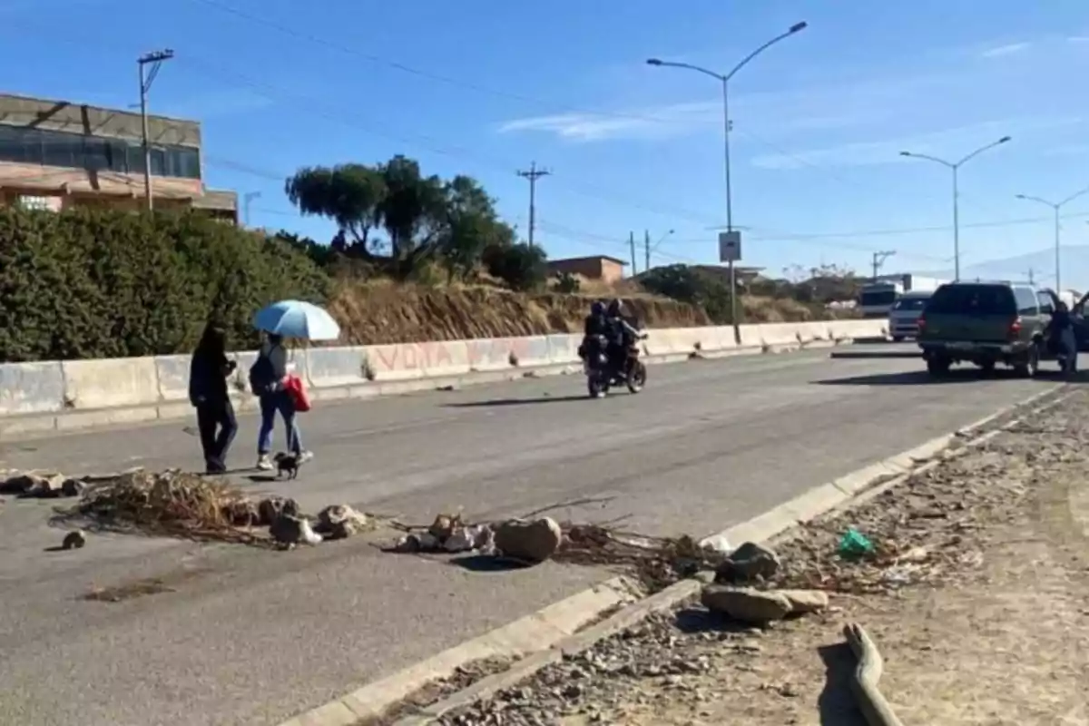 Personas cruzan una avenida con escombros y ramas en el suelo, una de ellas lleva una sombrilla azul, mientras pasan motociclistas y autos bajo un cielo despejado. Personas cruzan una avenida con escombros y ramas en el suelo, una de ellas lleva una sombrilla azul, mientras pasan motociclistas y autos bajo un cielo despejado.