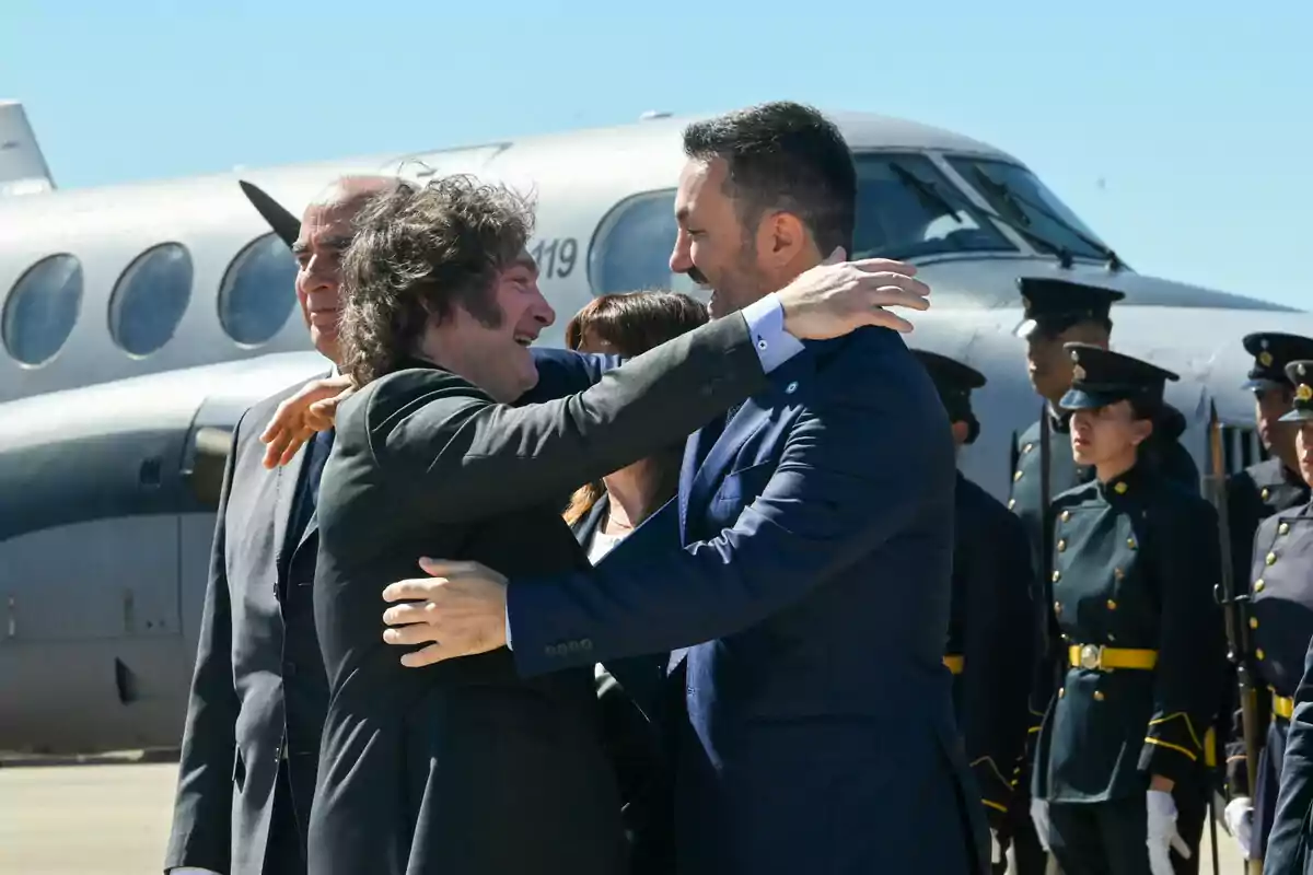 Two men smile and hug in front of an airplane while military personnel watch in the background. Two men smile and hug in front of an airplane while military personnel watch in the background.
