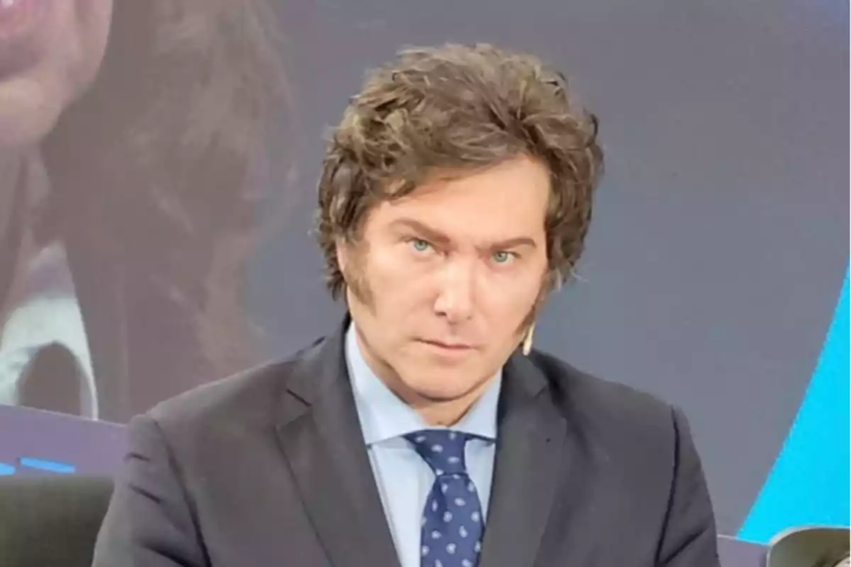 A man in a dark suit and a blue tie with white dots looking forward in a studio setting.