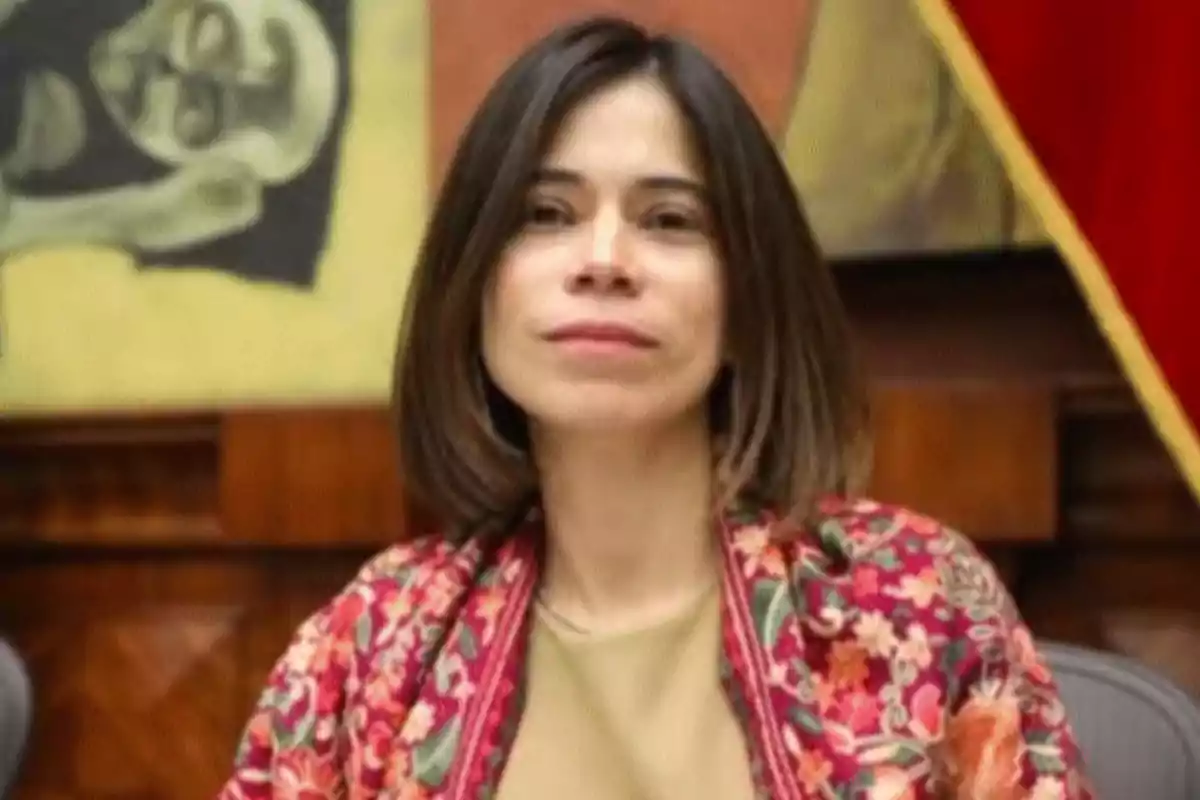 Short-haired, straight-haired woman with a serious expression wearing a colorful floral jacket, seated in front of an artistic background and a red flag.