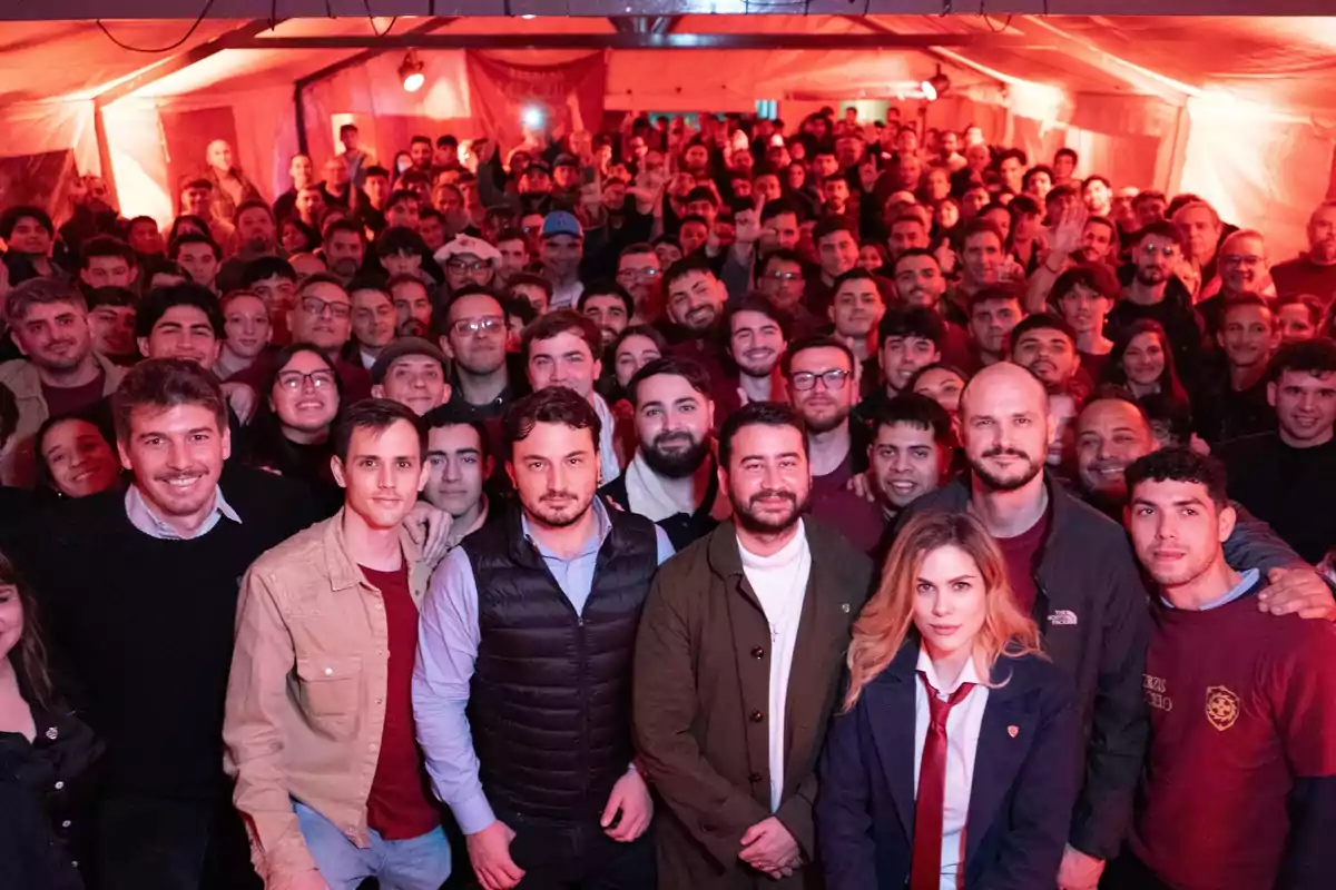 Un gran grupo de personas posando juntas dentro de una carpa iluminada con luces rojas, la mayoría sonríe y mira hacia la cámara