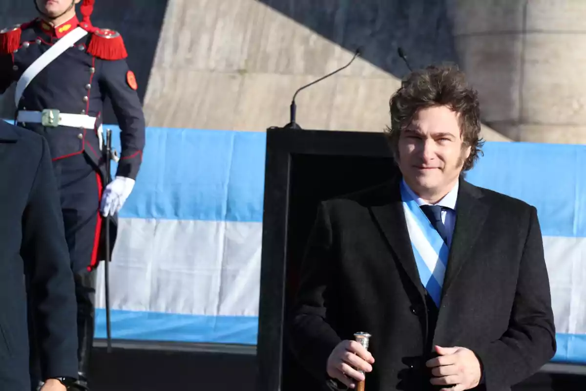 Un hombre con traje oscuro y corbata azul claro sostiene un bastón frente a una bandera argentina mientras un guardia con uniforme militar está de pie al fondo.