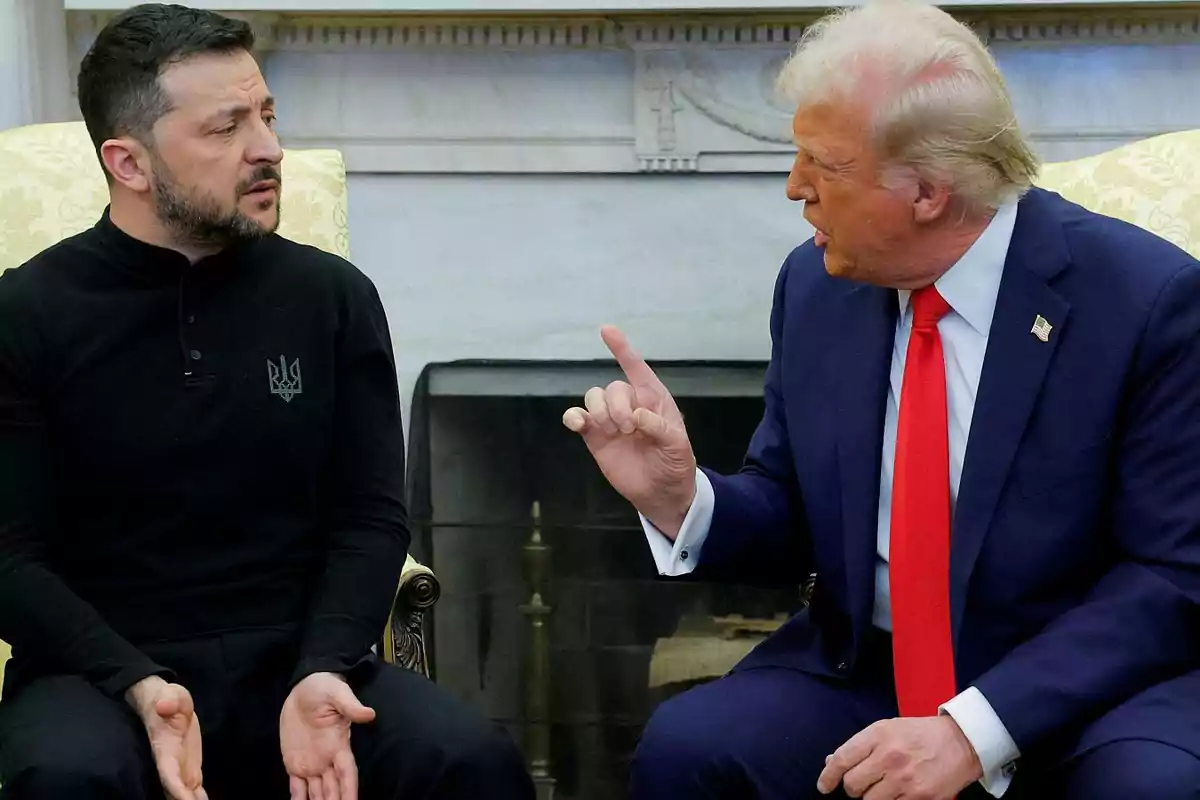 Two men sitting in a room, one in a dark suit and red tie gesturing with his hand, while the other, in a black sweater, watches him intently.