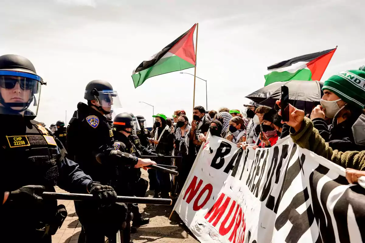 Policías con equipo antidisturbios frente a manifestantes que sostienen pancartas y banderas palestinas.