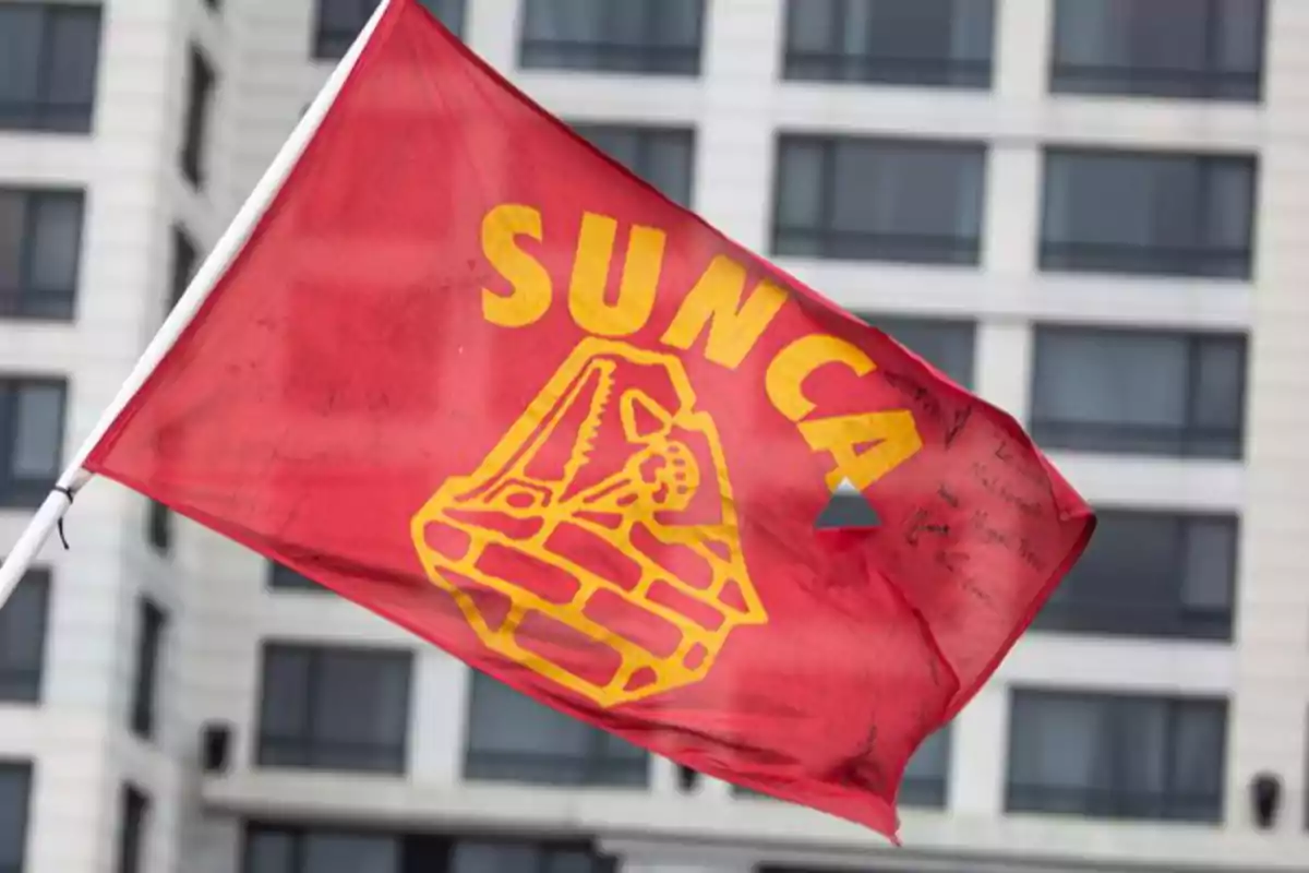 Bandera roja con el logo amarillo del sindicato SUNCA ondeando frente a un edificio moderno Bandera roja con el logo amarillo del sindicato SUNCA ondeando frente a un edificio moderno