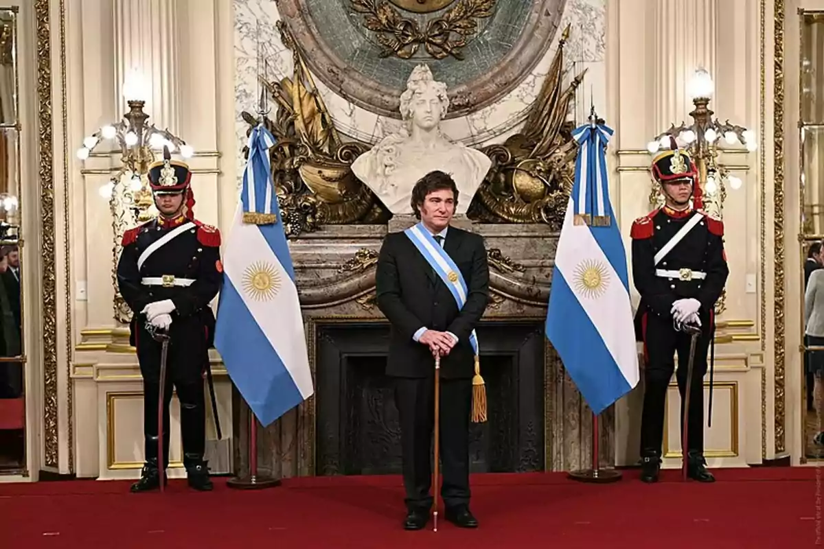 Una persona con banda presidencial está de pie entre dos guardias con uniformes ceremoniales y banderas argentinas en un salón decorado elegantemente.