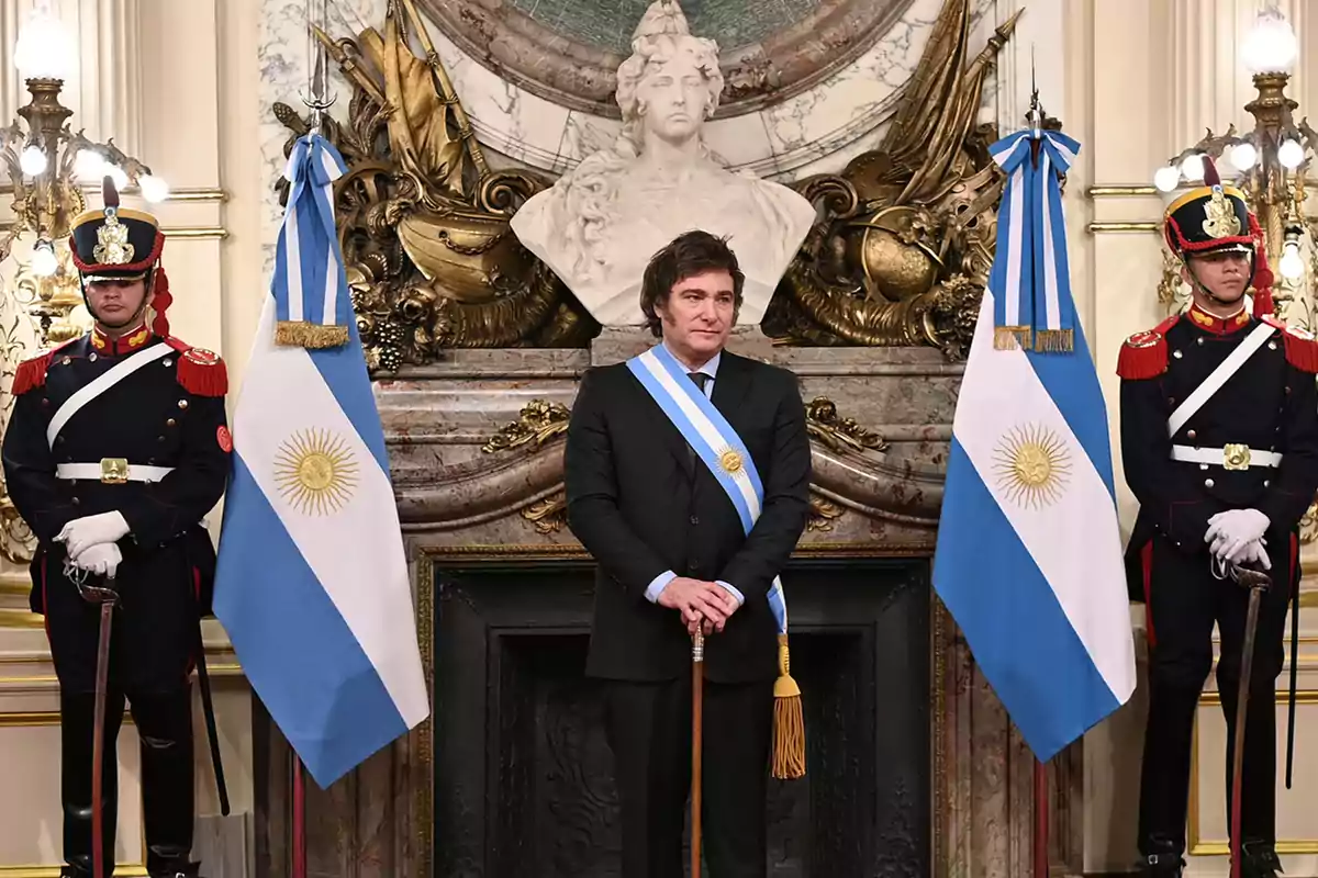 Un hombre con banda presidencial y bastón de mando está de pie entre dos banderas argentinas y dos guardias en uniforme ceremonial en un entorno elegante.