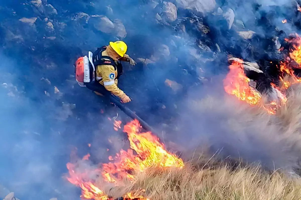 Un bombero con casco amarillo combate un incendio forestal entre humo y llamas en una zona de pastizales y rocas