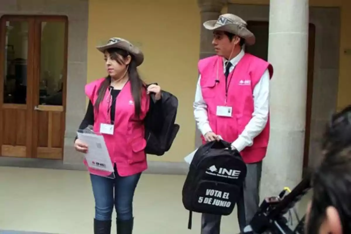 Two people with pink vests and hats hold INE backpacks in an indoor space. Two people with pink vests and hats hold INE backpacks in an indoor space.