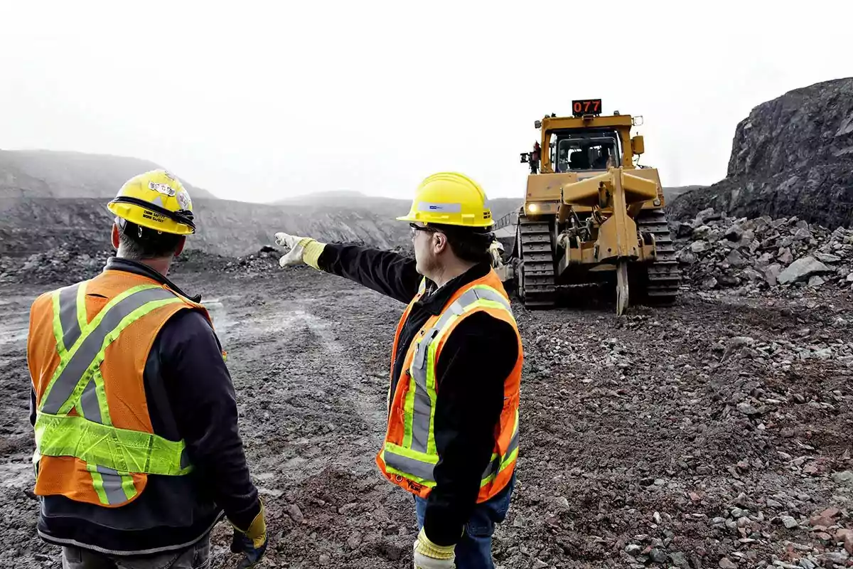 Dos trabajadores de la construcción con chalecos y cascos observan y señalan una excavadora en un sitio de trabajo rocoso.