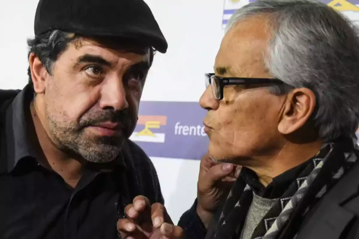 Two men are talking closely in an indoor setting, one wearing a beret and the other wearing glasses and a scarf, both with serious expressions.
