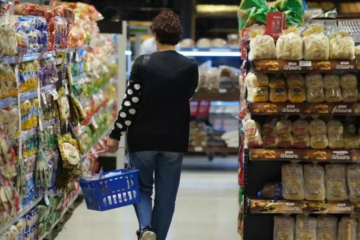 Una persona caminando por el pasillo de un supermercado con una canasta azul en la mano, rodeada de estantes llenos de productos.