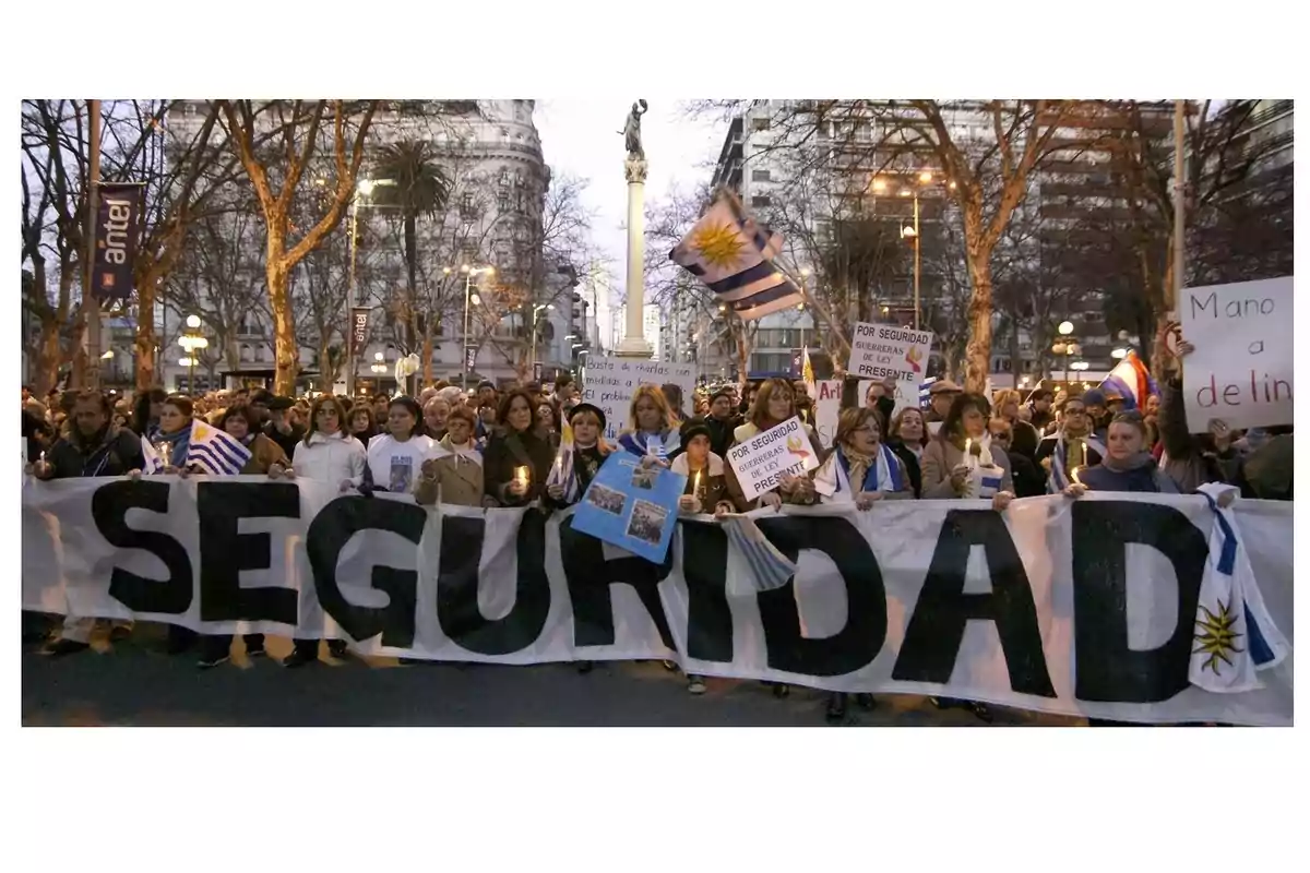 Un grupo de personas participa en una manifestación en una calle arbolada, sosteniendo una pancarta grande que dice Un grupo de personas participa en una manifestación en una calle arbolada, sosteniendo una pancarta grande que dice