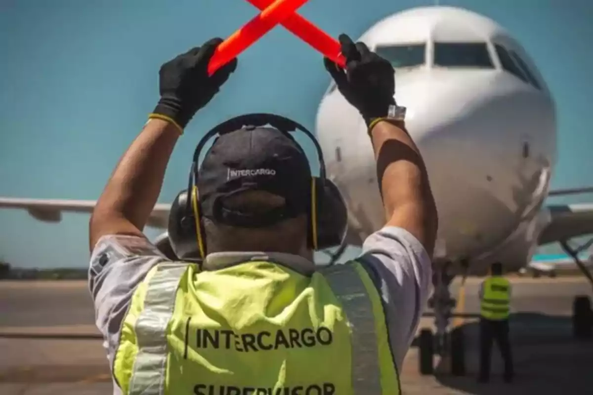 Un trabajador aeroportuario con chaleco amarillo y auriculares guía a un avión en la pista usando bastones de señalización rojos.