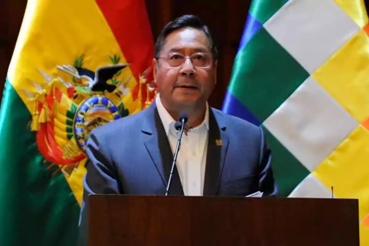 Man with glasses and a dark suit speaking at a podium with the flags of Bolivia and the Wiphala in the background Man with glasses and a dark suit speaking at a podium with the flags of Bolivia and the Wiphala in the background