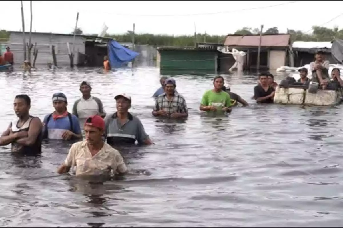 Un grupo de personas camina por una zona inundada con agua hasta la cintura, con casas y estructuras en el fondo parcialmente sumergidas.