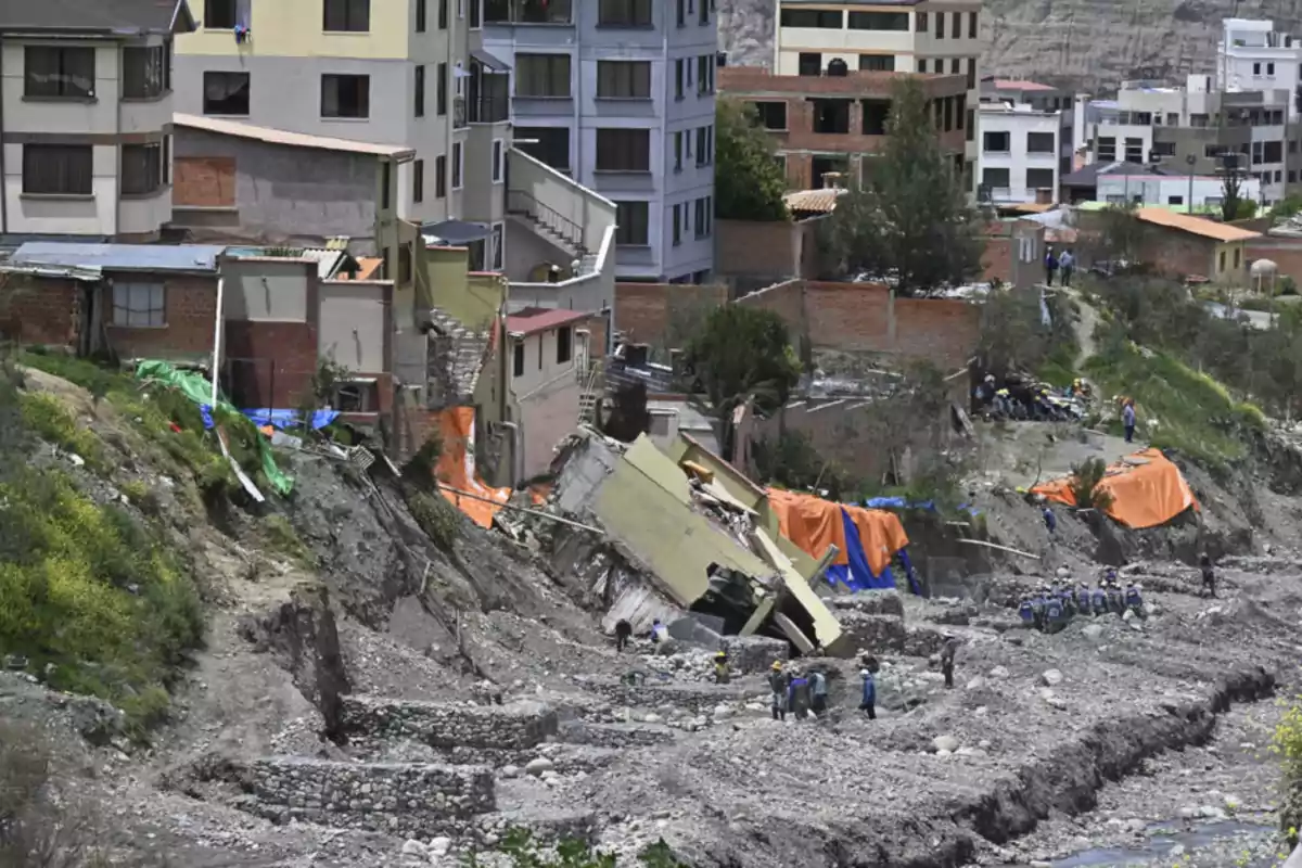 Edificios en una ladera con deslizamiento de tierra y estructuras colapsadas, mientras personas observan y trabajan en el área afectada.