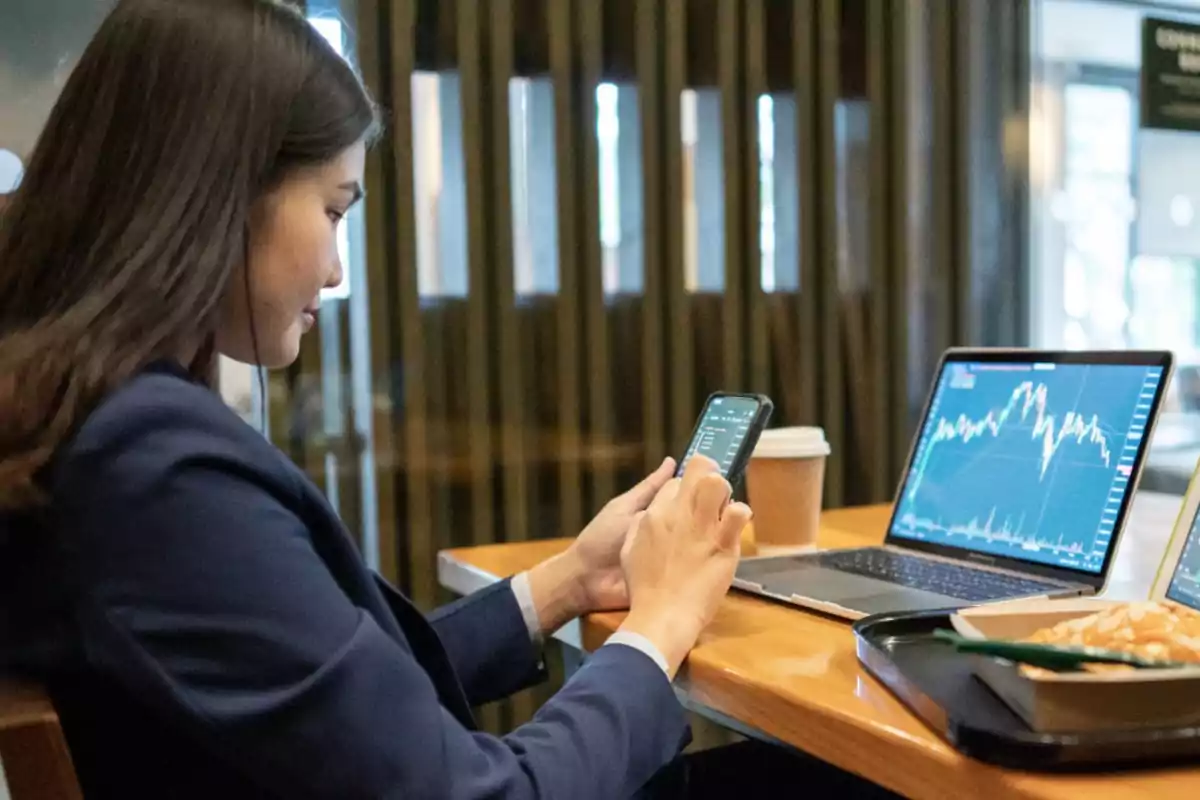 Mujer en una cafetería usando un teléfono móvil con gráficos financieros en la pantalla de una laptop.