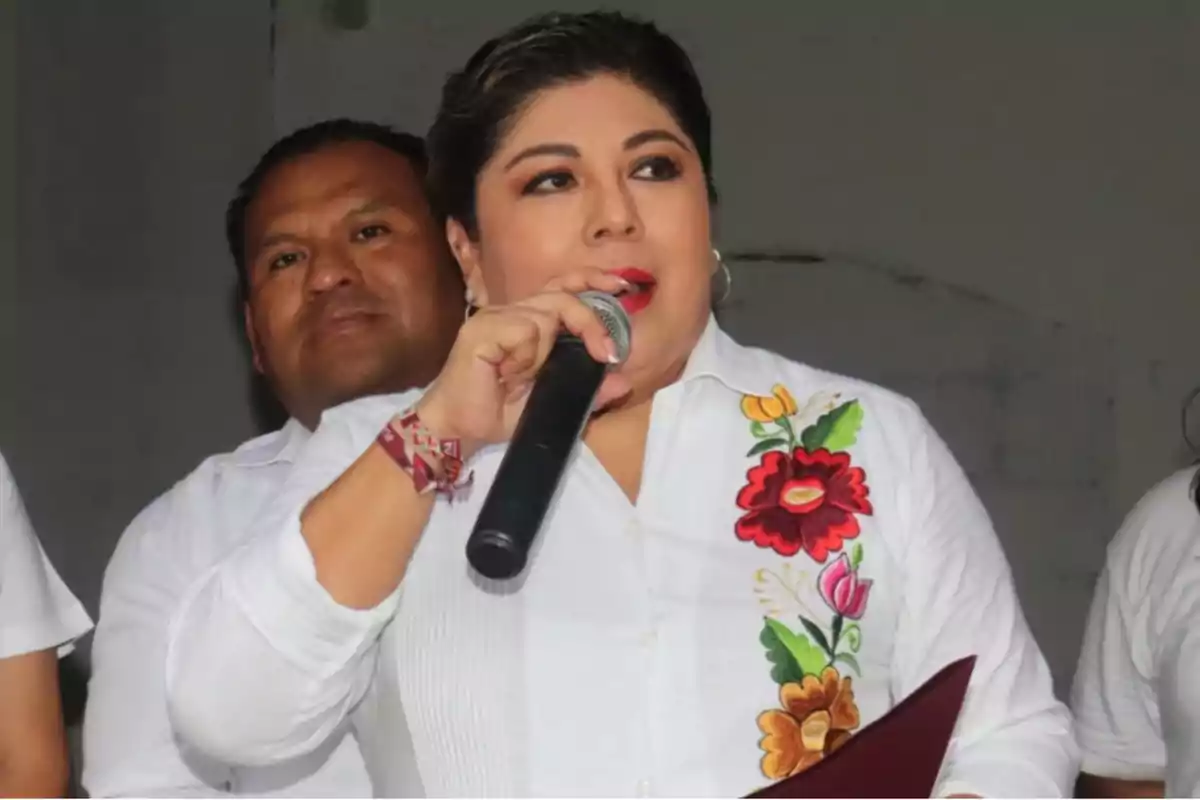 Una persona hablando por micrófono con una camisa blanca bordada con flores de colores. Una persona hablando por micrófono con una camisa blanca bordada con flores de colores.
