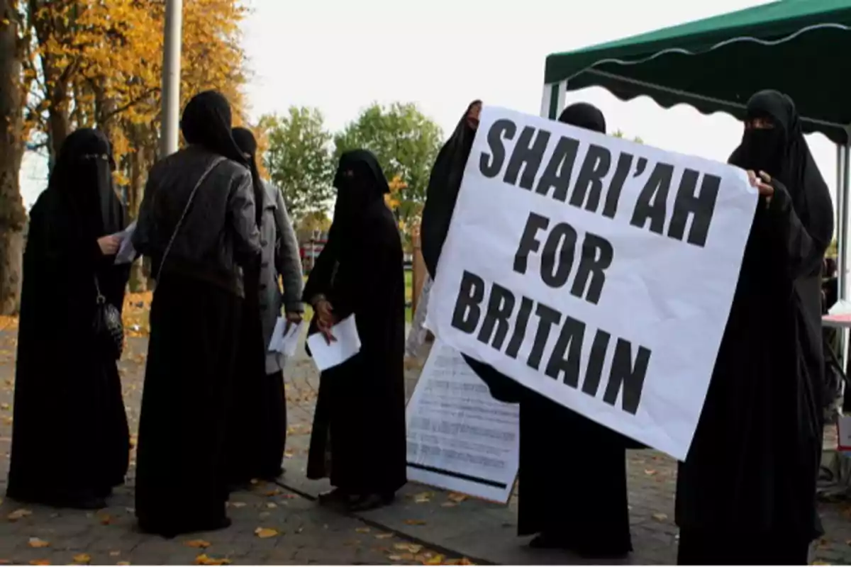 Un grupo de mujeres vestidas con niqabs sostiene un cartel que dice "Shari'ah for Britain" en un entorno al aire libre con árboles y hojas caídas.