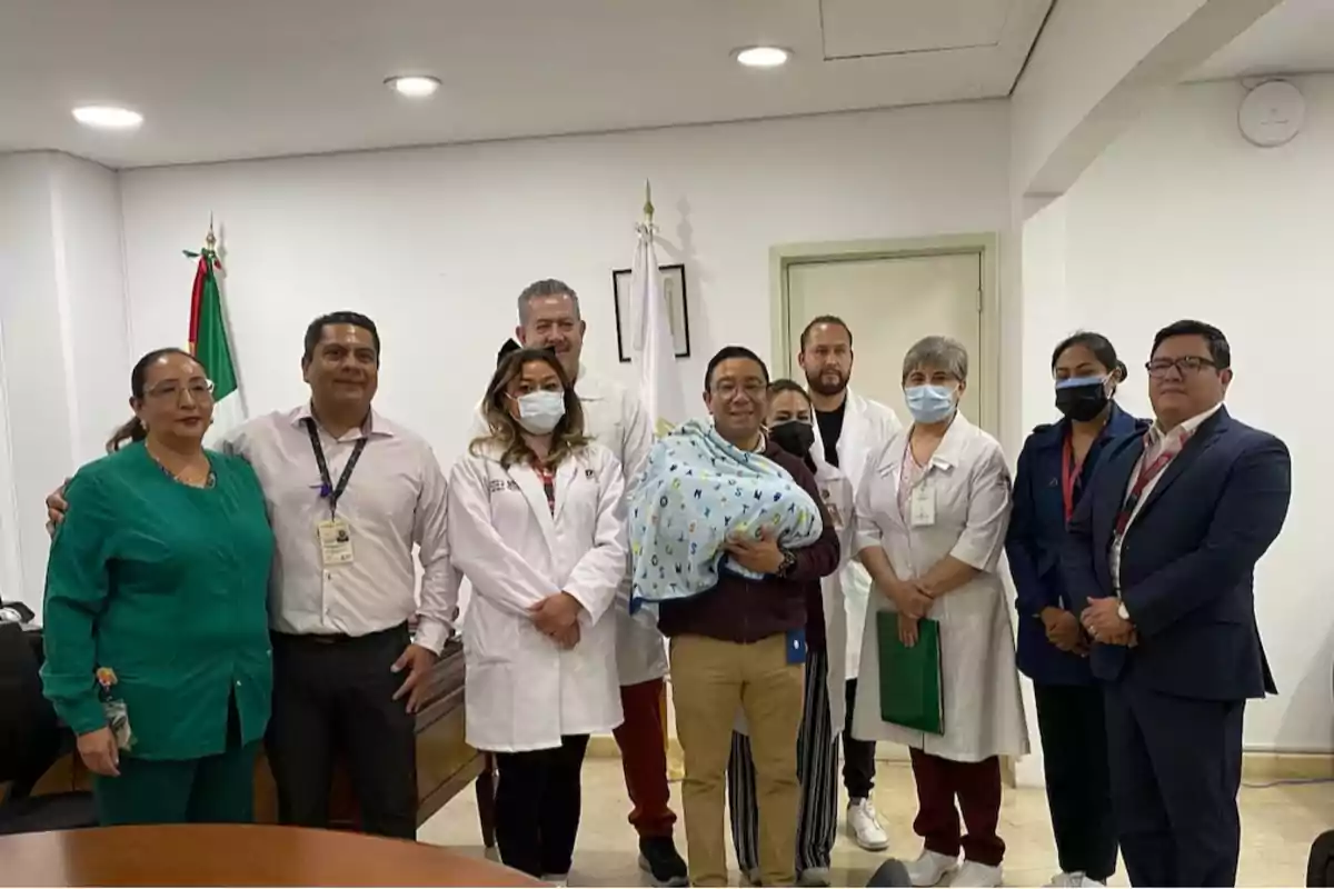 A group of people, some in medical gowns, pose for a photo in a room with a Mexican flag in the background; one person is holding a baby wrapped in a blanket. A group of people, some in medical gowns, pose for a photo in a room with a Mexican flag in the background; one person is holding a baby wrapped in a blanket.