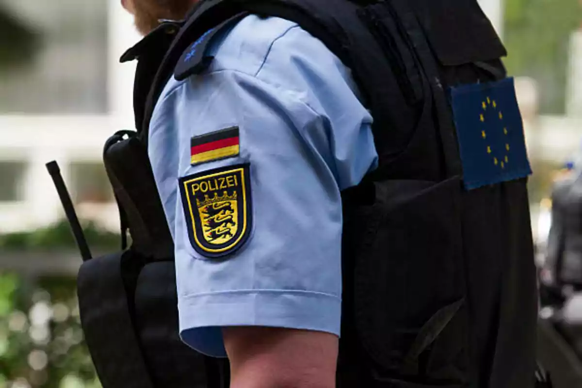 Un oficial de policía con uniforme azul y chaleco negro lleva un parche de la bandera alemana y un escudo que dice "Polizei" en el brazo izquierdo.